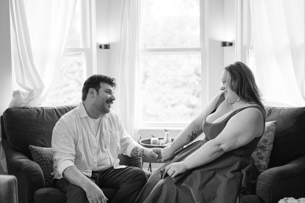 Black and white photo of a couple sitting on a couch, smiling and talking in a bright living room.