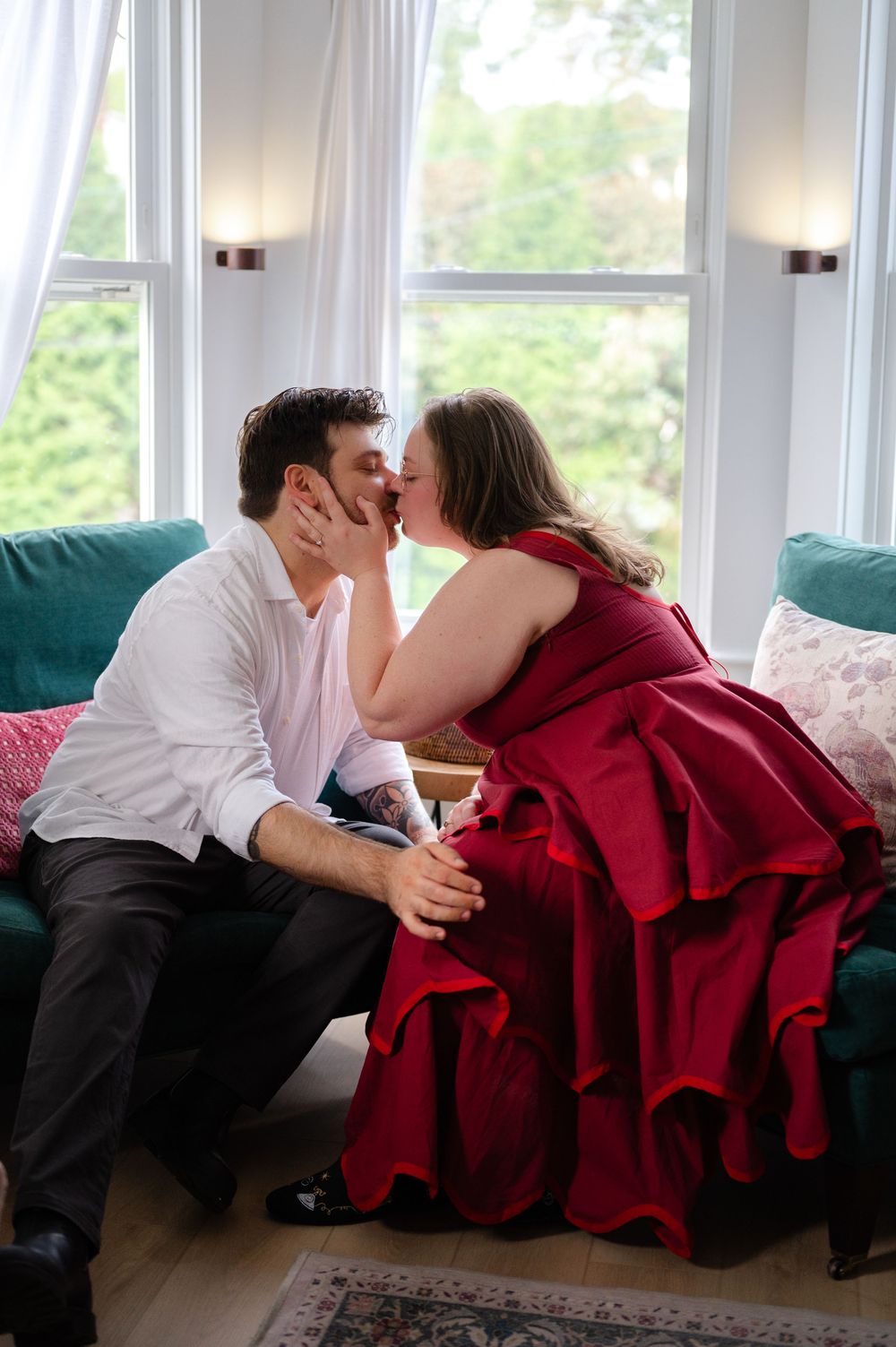 A couple sharing a romantic kiss on a teal sofa, woman in red gown, man in white shirt, near bright bay windows.
