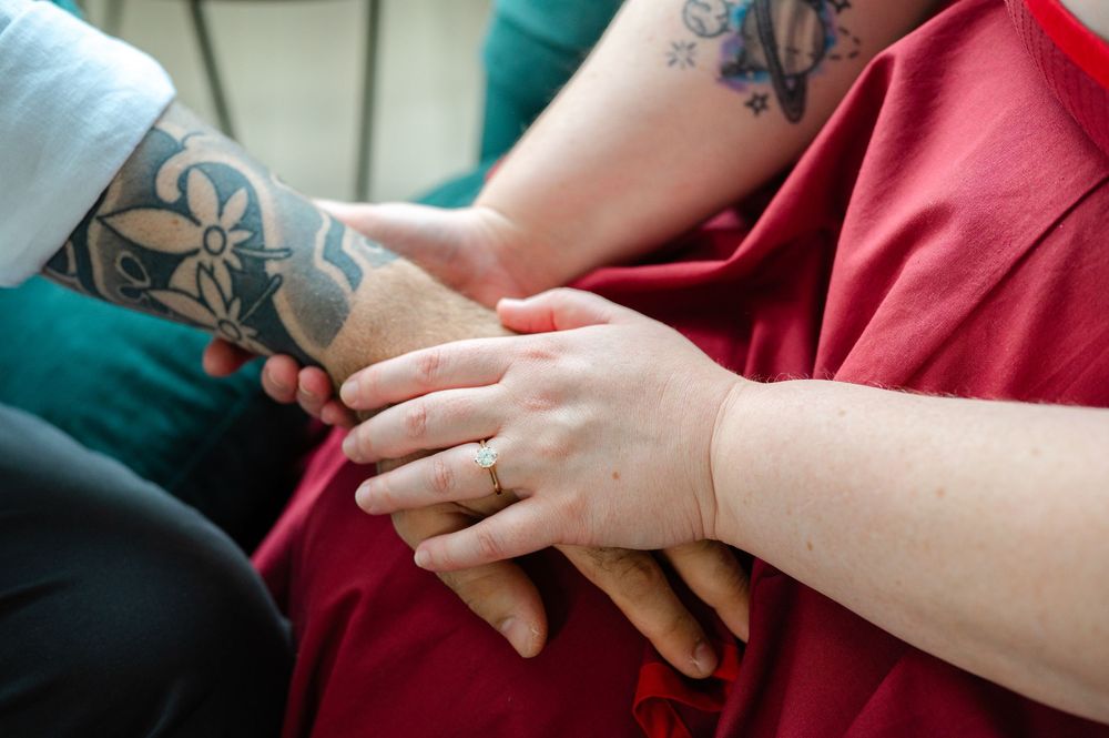 Two people holding hands, one with floral tattoos on their arm, wearing a red shirt and a gold ring.