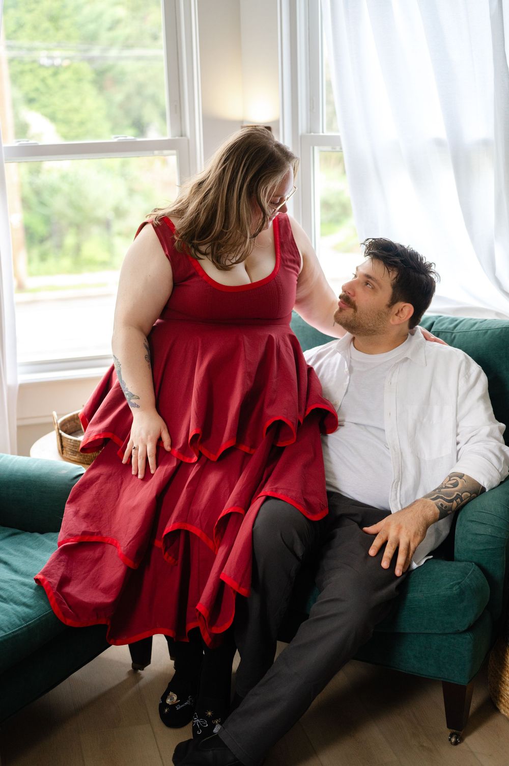 A plus-size woman in a red ruffled dress stands beside a man in white sitting on a green sofa.