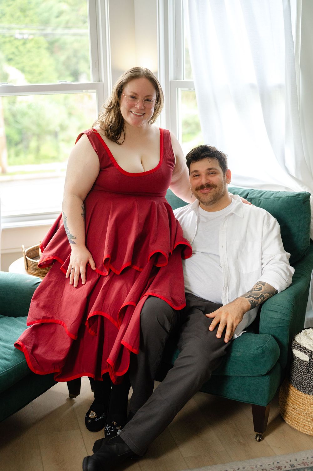 A woman in a red dress and a tattooed man in white shirt smile together on a green sofa by a window.
