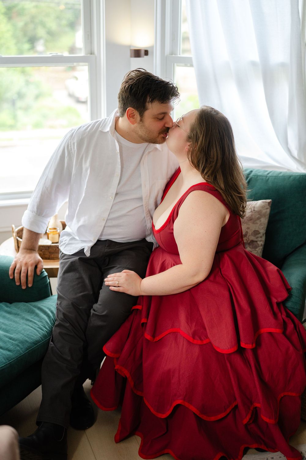 Couple sharing a kiss on a green sofa, woman in red ruffled gown and man in white shirt.