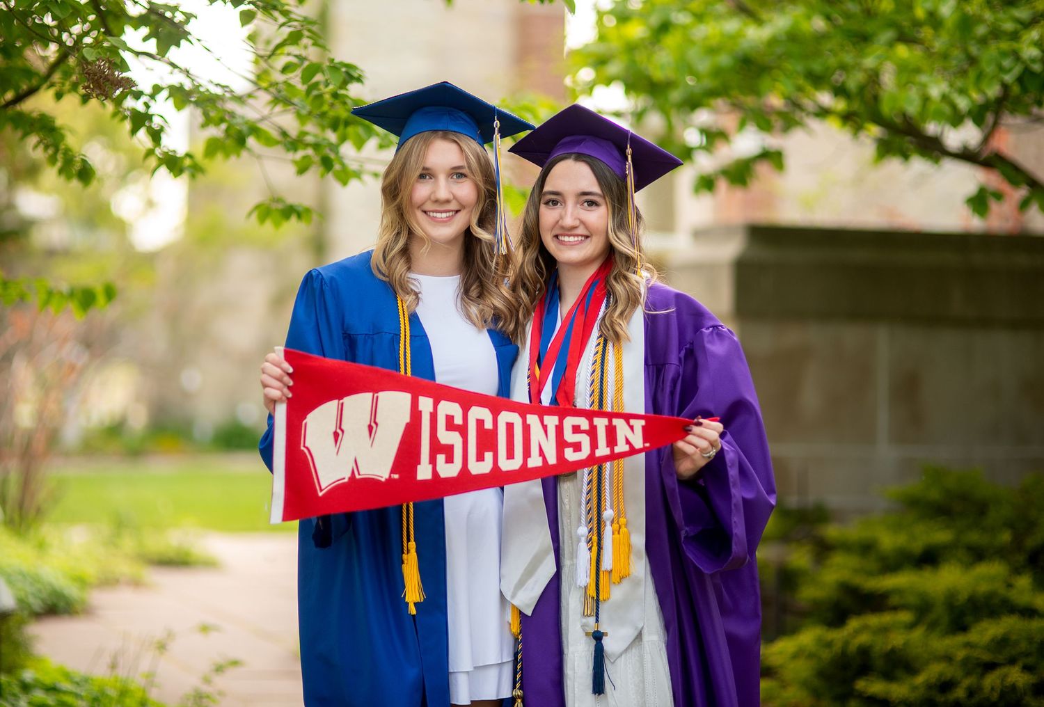 Two female graduates in caps and gowns holding a Wisconsin Badgers pennant outdoors.