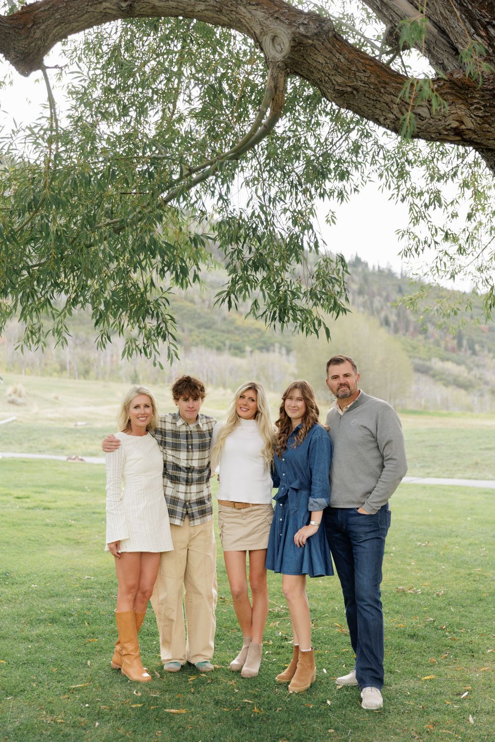 Family of five poses under large tree in green park with mountain landscape in background.