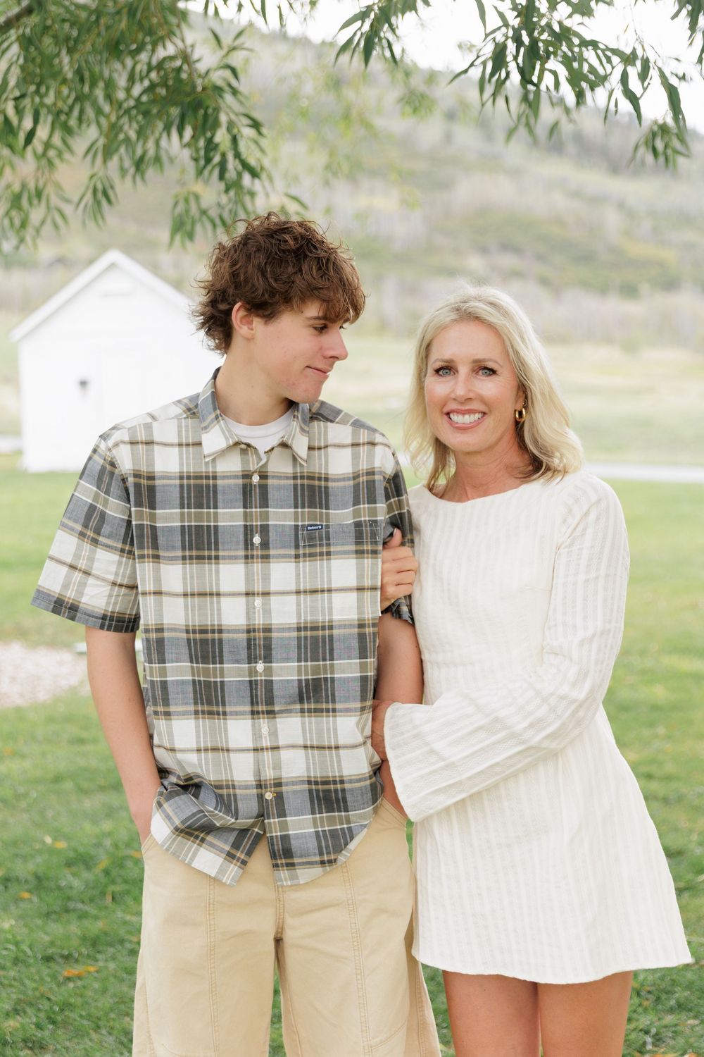 A teenage boy in plaid shirt and blonde woman in white dress pose together outdoors in a green meadow setting.