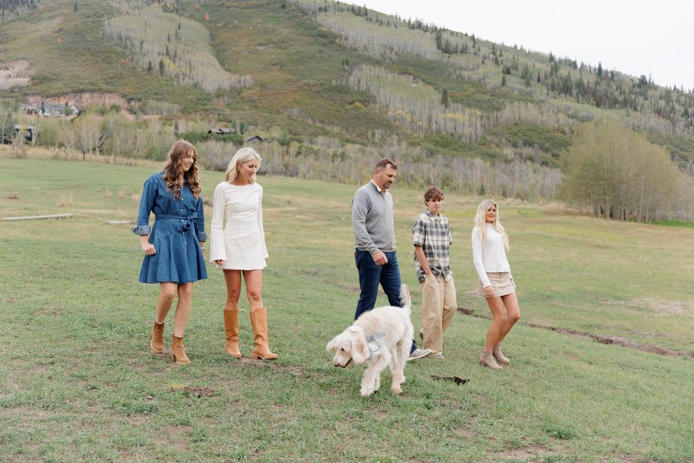 A family of five walks with their white fluffy dog across a scenic mountain meadow in autumn.