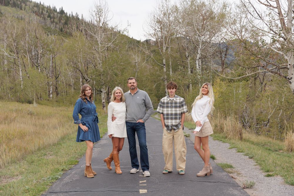 A family of five poses together on a paved path surrounded by autumn trees in a mountain setting.