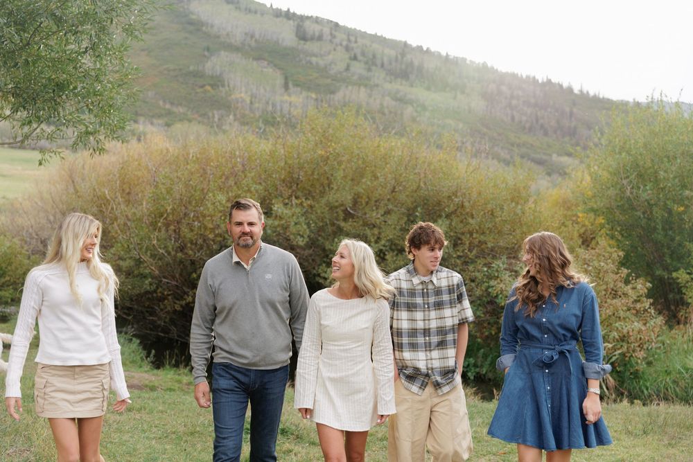 Family of five walks together through green meadow with rolling hills and shrubs in background.