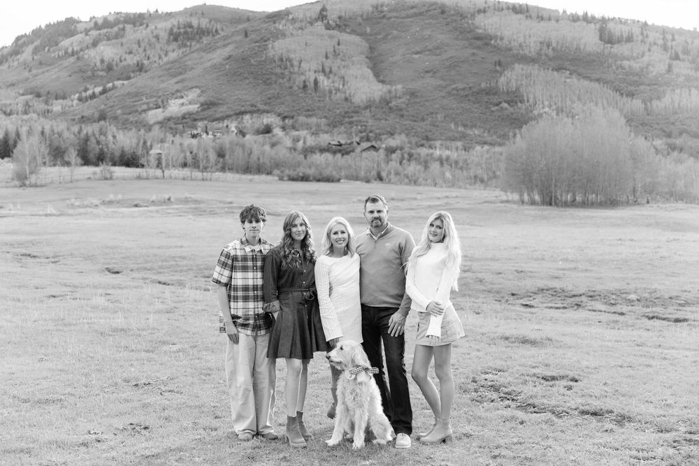 Black and white photo of family of five with dog posing in open field with mountain backdrop.