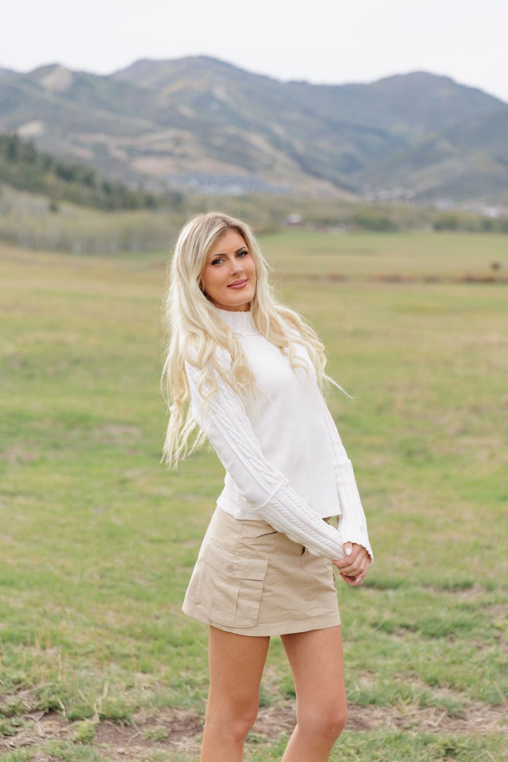 Blonde woman in white sweater and tan skirt poses in mountain meadow with hills in background.