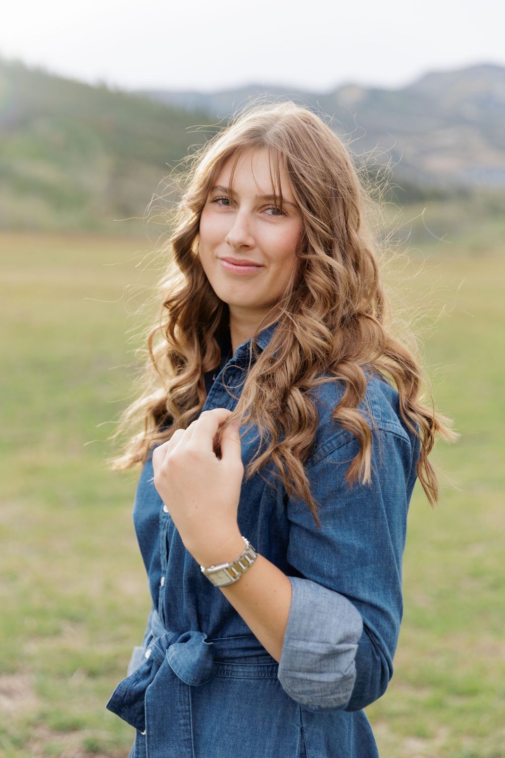 Young woman with curly auburn hair in denim dress smiles in mountain meadow setting.