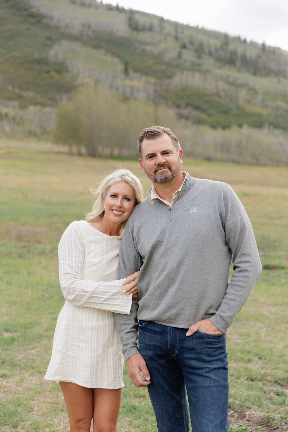 Smiling couple poses together in green mountain meadow, woman in white dress, man in gray pullover.