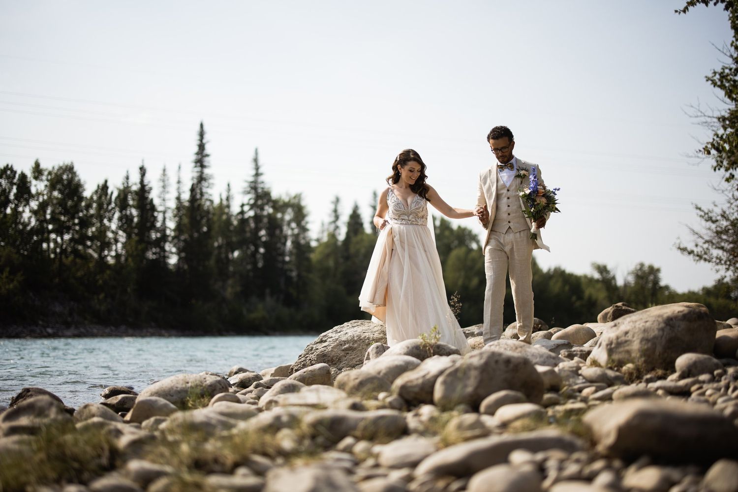 Bride and groom holding hands on a rocky riverbank with a line of evergreen trees