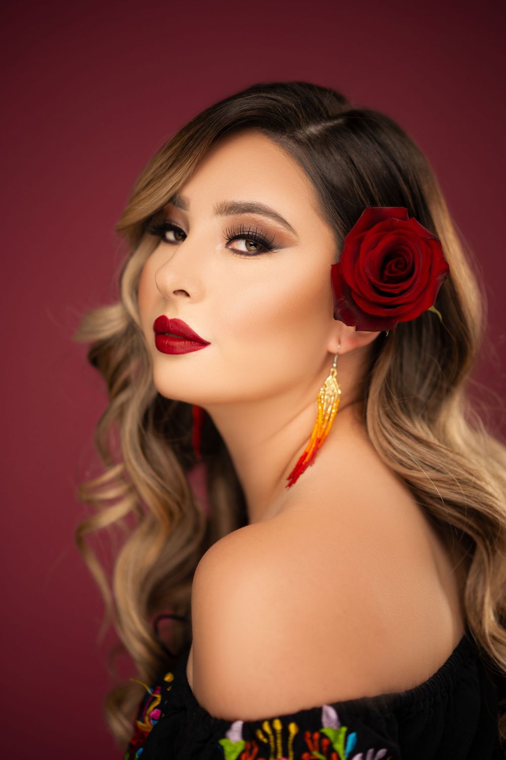 Glamorous beauty portrait of a woman wearing red lipstick, gold earrings, and a red rose in her hair against a burgundy background.