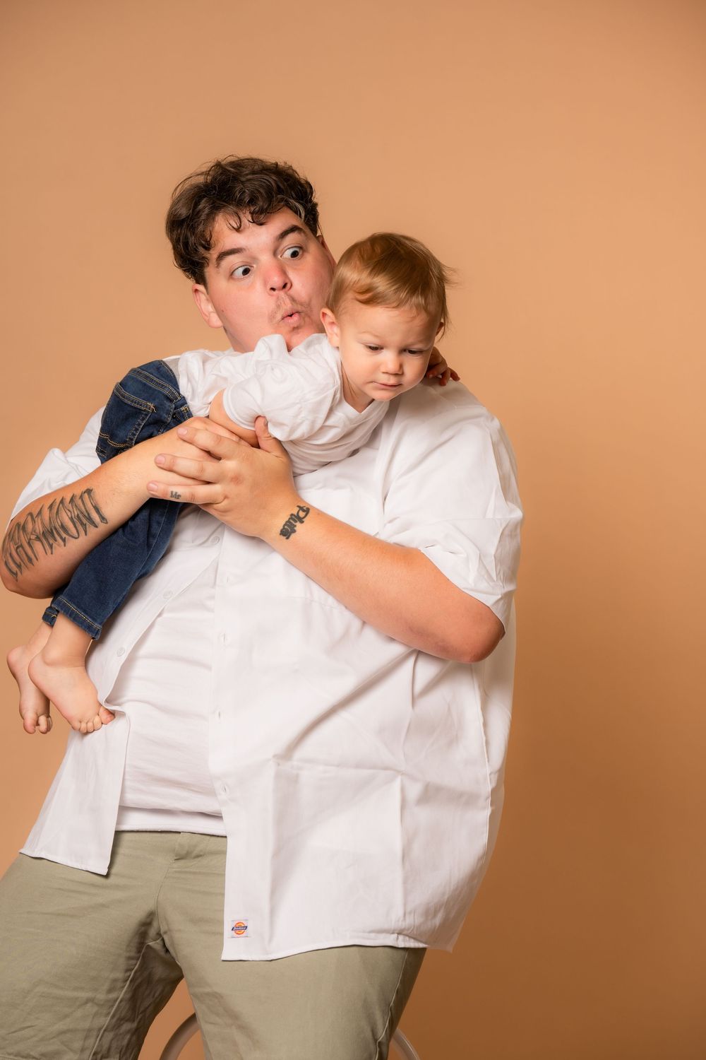 Playful studio portrait of a man holding a young child against a warm peach-colored background.