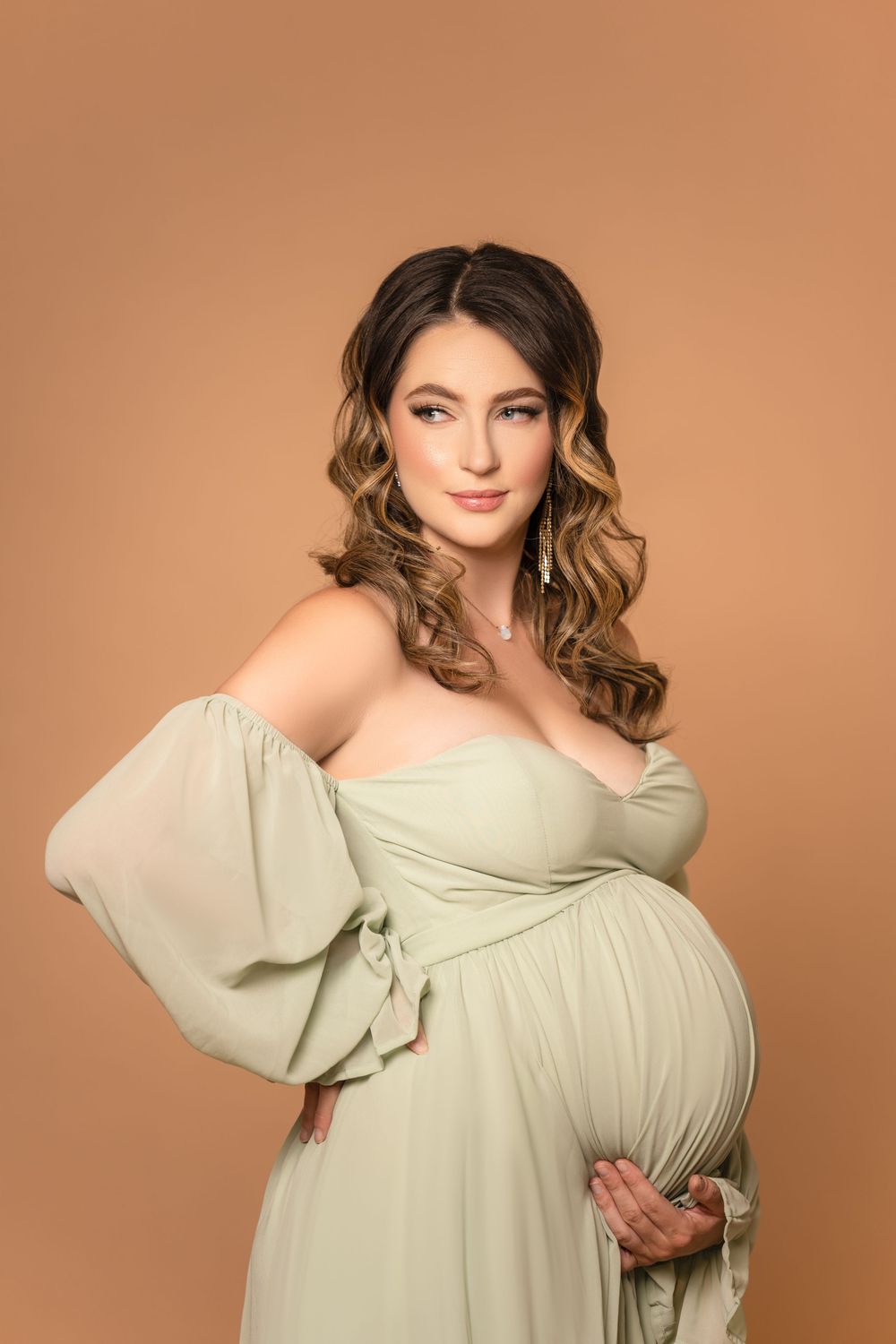 Pregnant woman in a pale green off-shoulder dress gently holding her belly against a warm beige studio background.