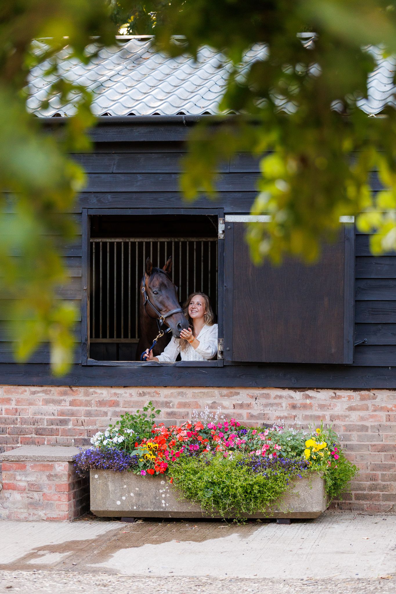 Woman with long blonde hair in a navy blue gown stands beside a bay horse in a bridle, with green hedges and a brick building behind.