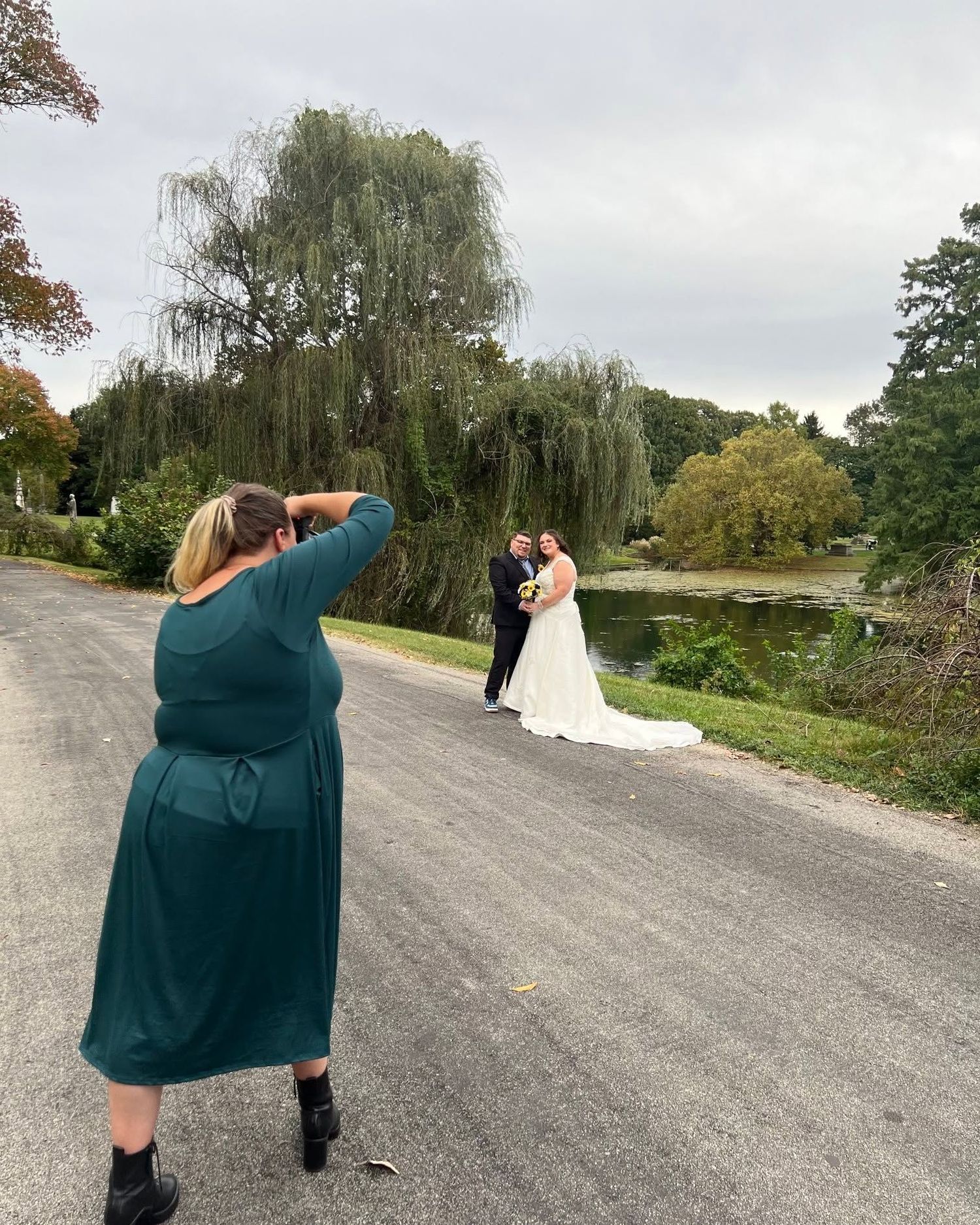 A photographer in a green dress photographs a bride and groom on a country path beside a pond and weeping willow trees.