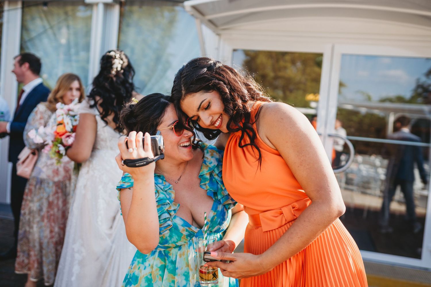 Two bridesmaids in colorful dresses laughing and looking at a phone at an outdoor wedding reception at Morden House Registry Office London