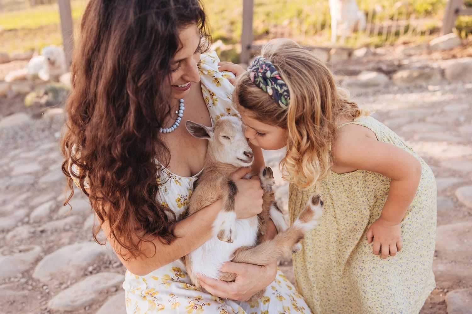 A smiling mother and her young daughter in a yellow floral dress gently cuddle a baby goat on a sunny farm.