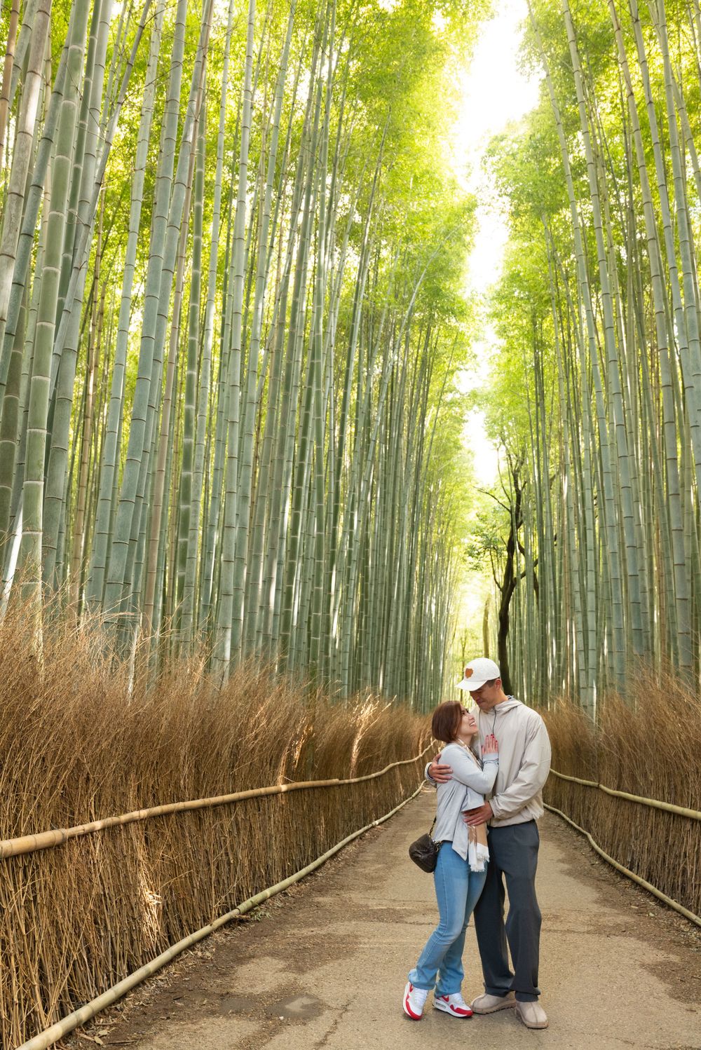 Couple embracing on a stone path lined with tall green bamboo stalks and woven fences in the Arashiyama Bamboo Grove at sunset.