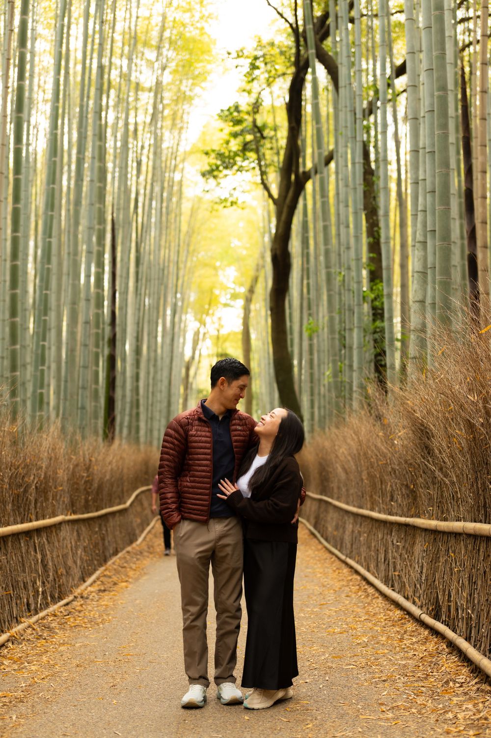 Couple embracing and smiling on a path lined with wooden fencing, dry grass, and tall green bamboo trees in a peaceful forest.