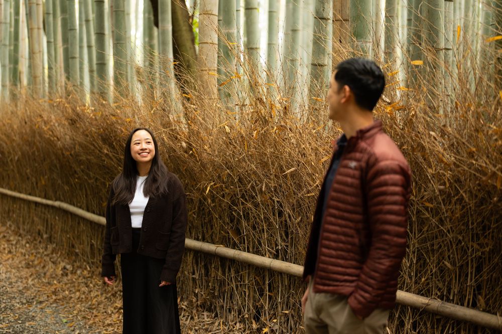 Smiling woman in black and man in a red puffer jacket stand beside a wooden railing in a tall bamboo grove.