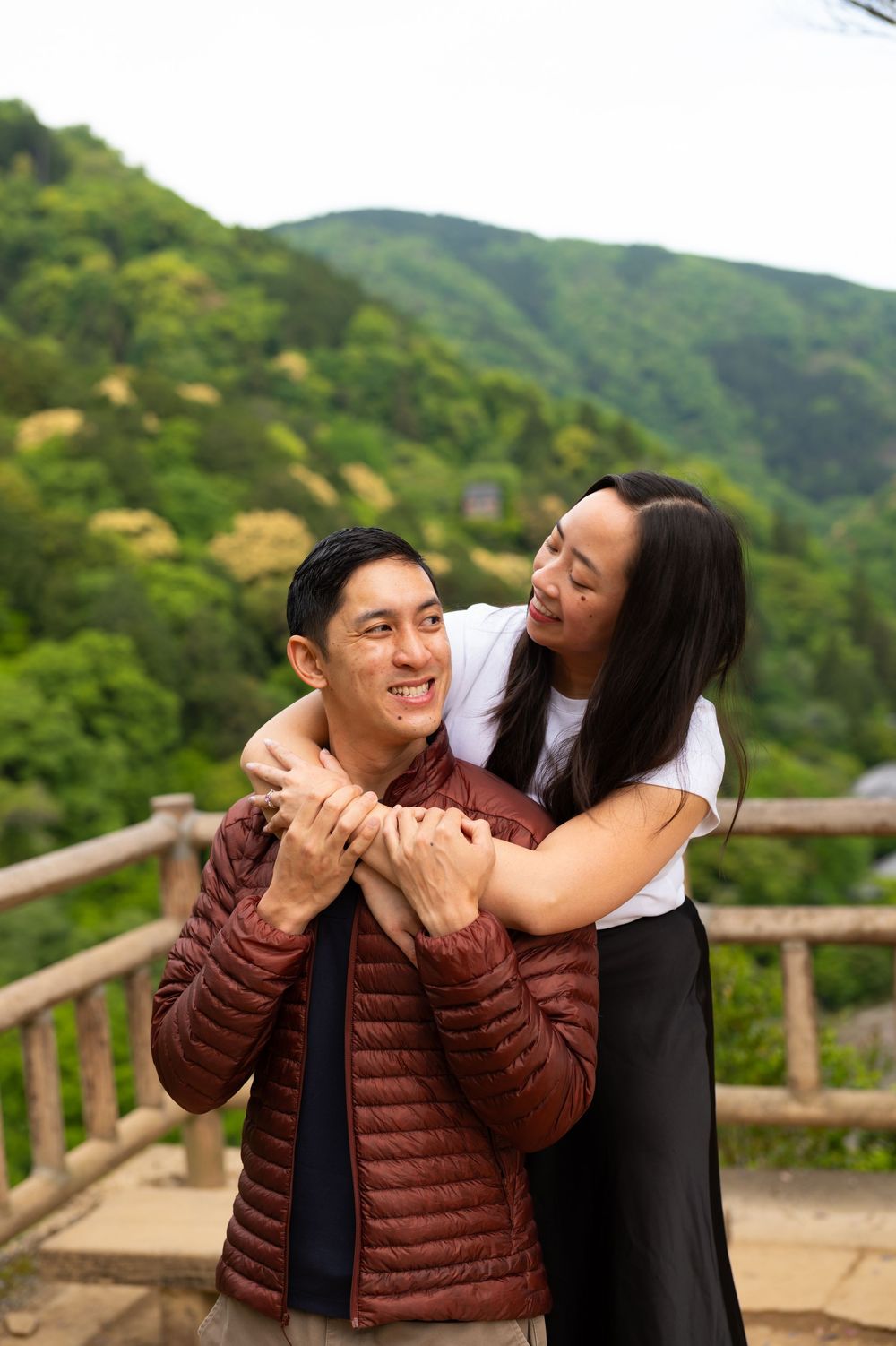 Smiling couple embraces on a wooden overlook, a woman hugging a man in a red puffer jacket from behind with green forested mountains behind.