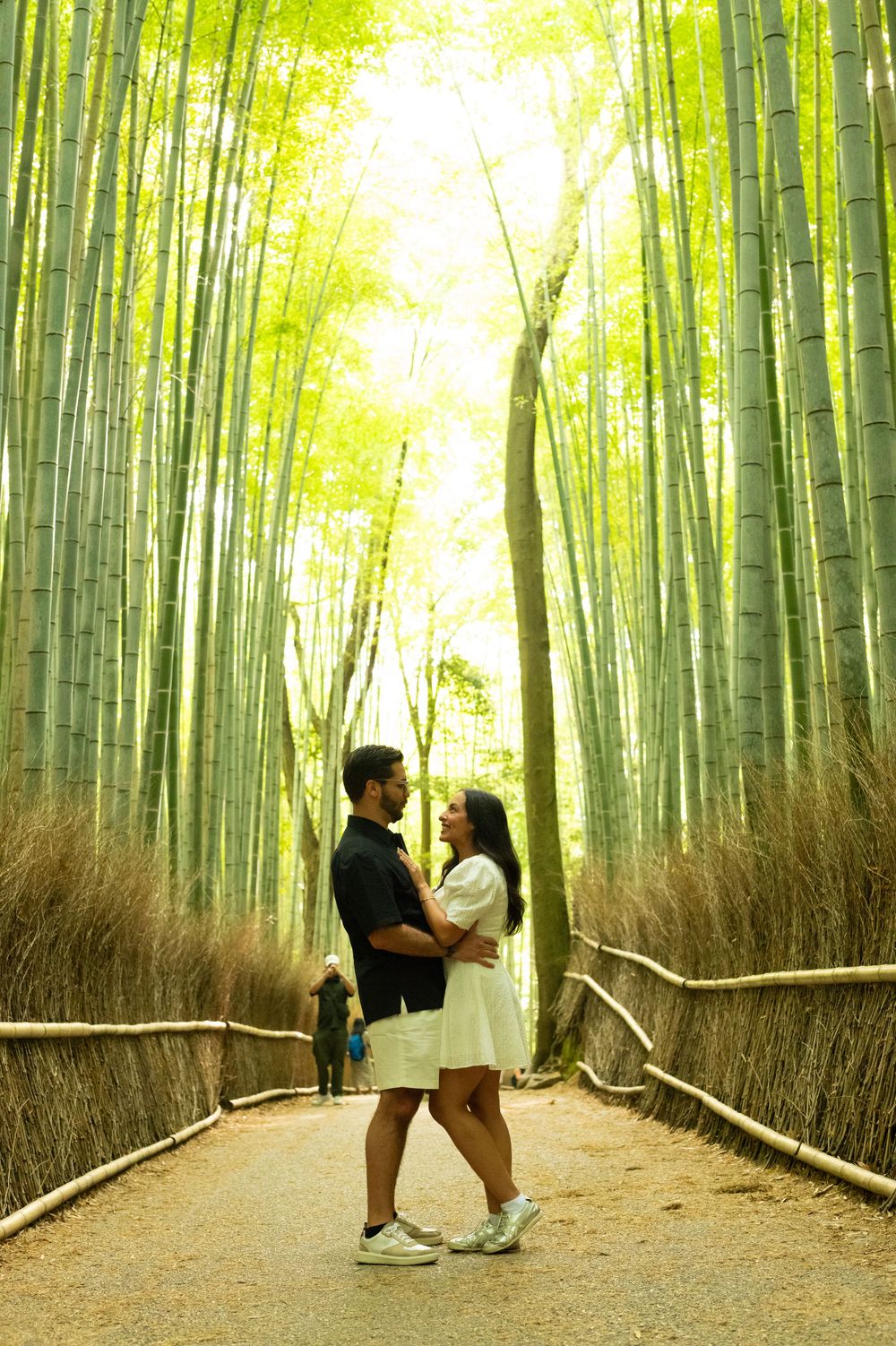 Couple embracing on a path through a sunlit bamboo grove in Arashiyama, Kyoto, with tall green bamboo stalks and woven fences.