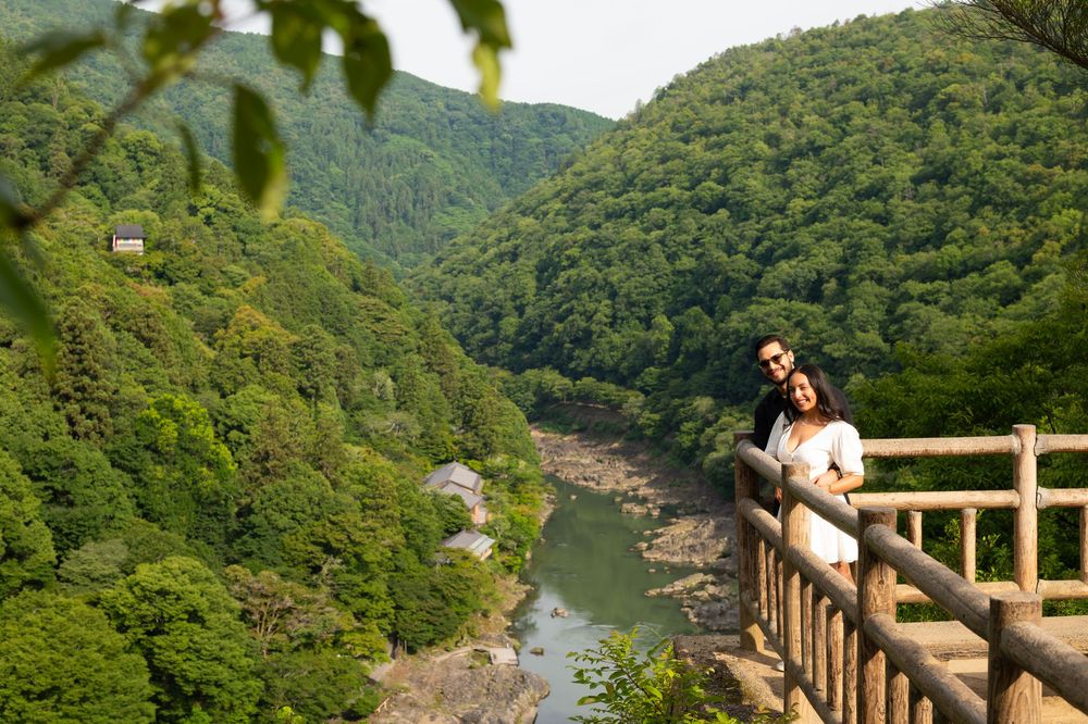 Couple embracing on a wooden observation deck overlooking a calm river and lush green forested mountains in a scenic valley.