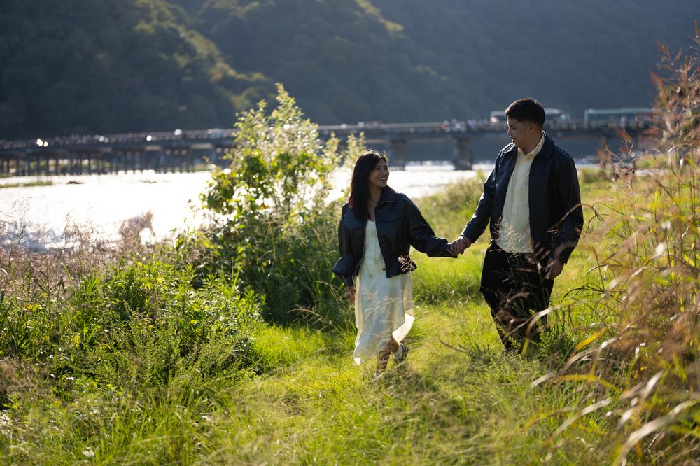 A young couple in dark jackets holds hands while walking through a sunlit grassy field beside a calm river with a bridge and forested hills.