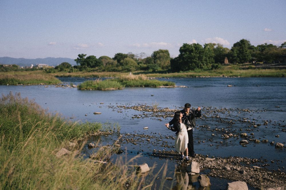 A couple in wedding attire poses on rocks in a shallow river, with grassy banks, green trees and distant hills under a clear blue sky.
