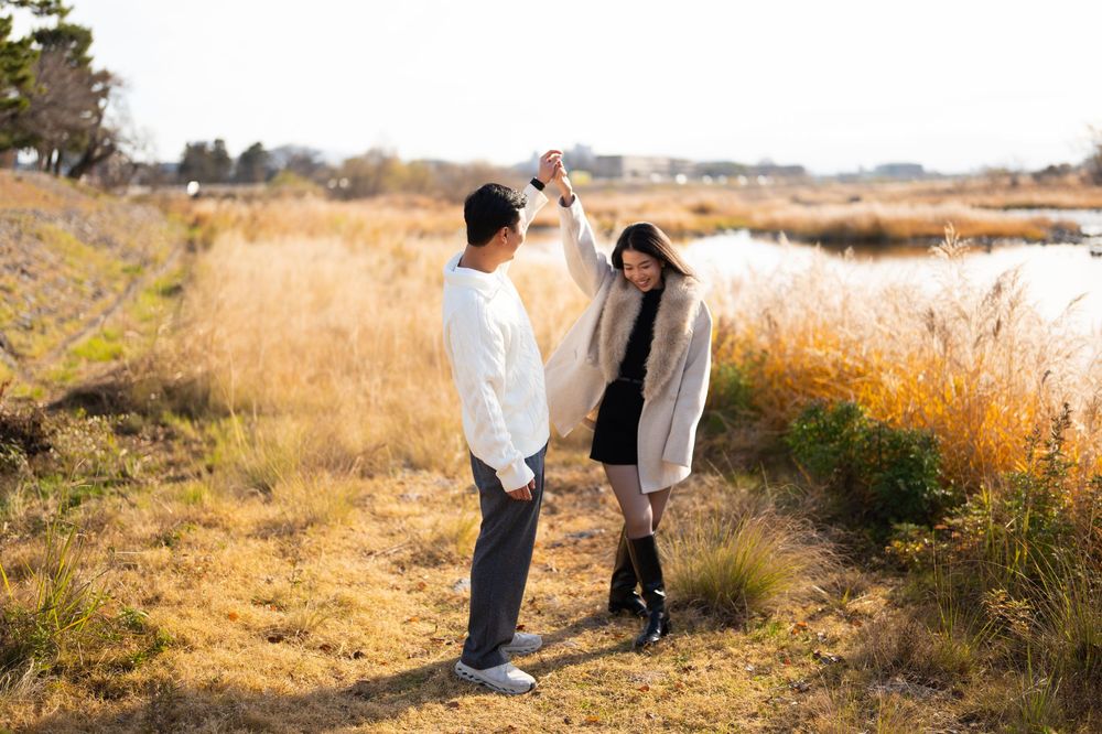 Couple dancing hand in hand on a grassy golden field beside a calm lake during a sunny outdoor engagement photo session.