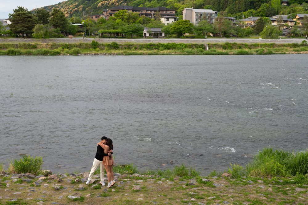 Couple embracing on a rocky riverbank beside a wide calm river, with houses and green forested hills in the background.