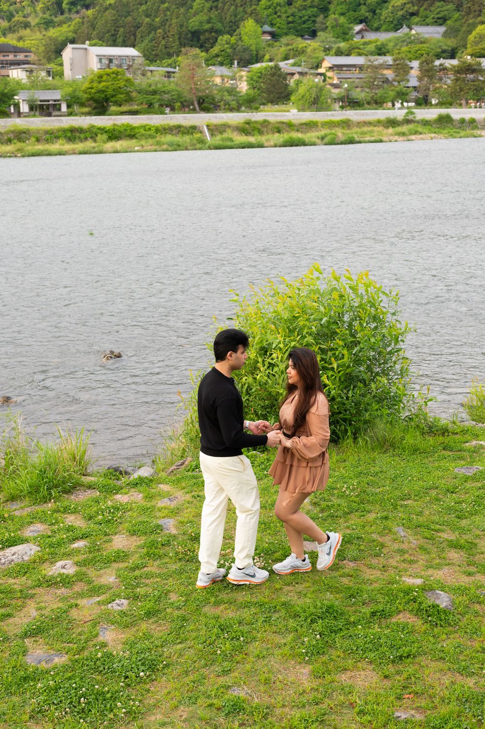 A couple holds hands facing each other on a grassy lakeside lawn with green shrubs, calm water, and village houses in the background.