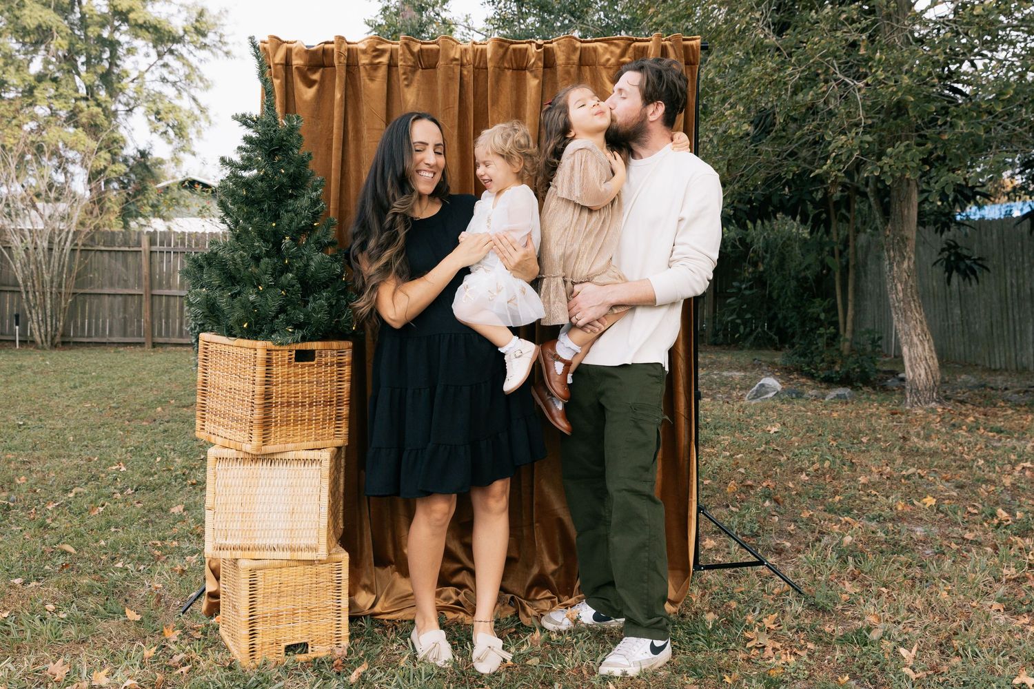 Parents hold two young daughters in front of a brown curtain backdrop in a backyard with wicker baskets and a small tree.