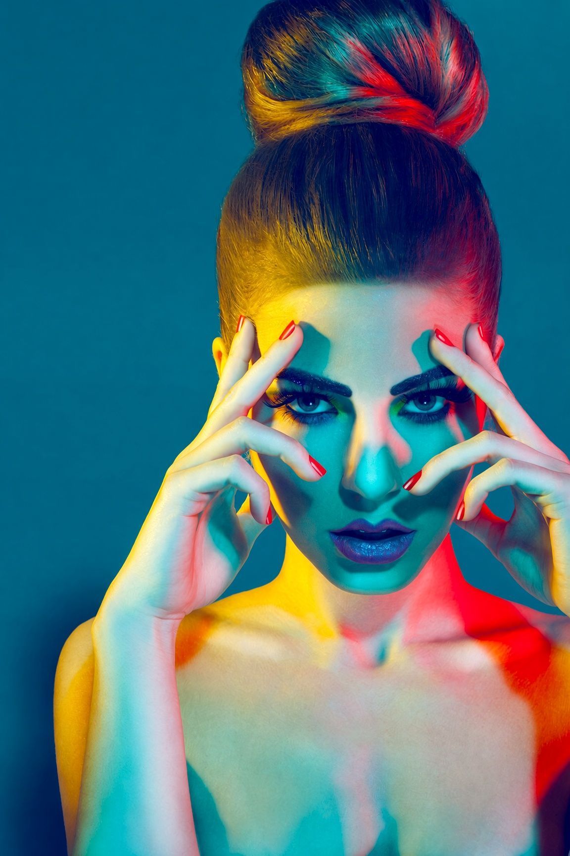 Fashion portrait of a woman with a high bun and red nails framing her face, lit by vivid teal, yellow and red studio lights.
