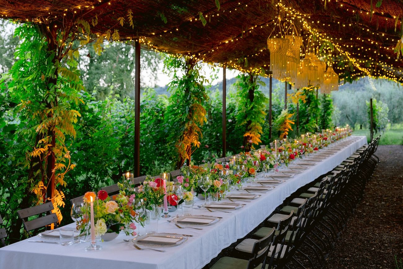 A colourful rehearsal dinner table with candles under a leaf canopy & rope chandeliers in a Lake Garda vineyard. Design by Solidea Events