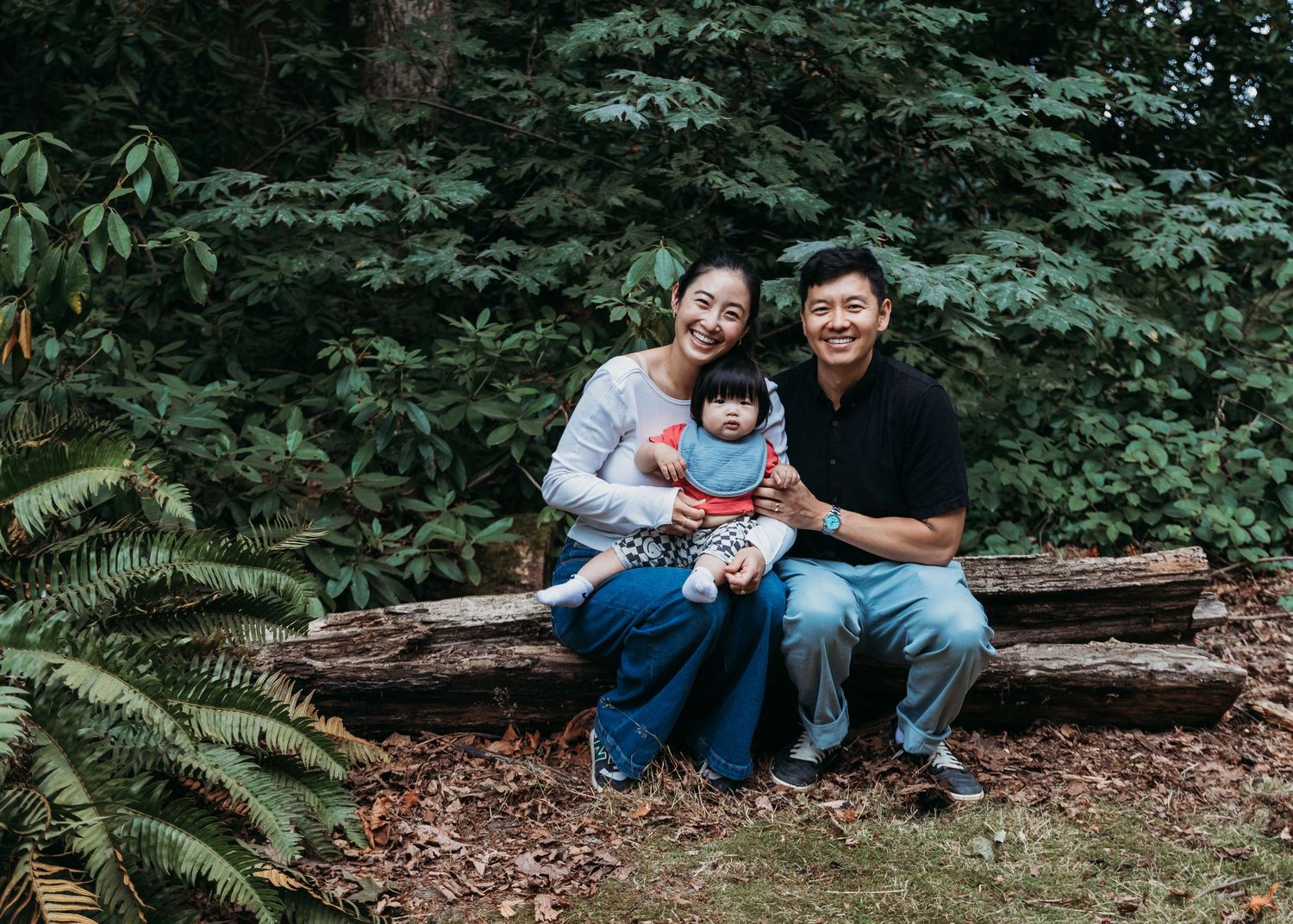 Smiling couple sits on a mossy log in a lush green forest holding their baby in a red outfit during an outdoor family portrait session.