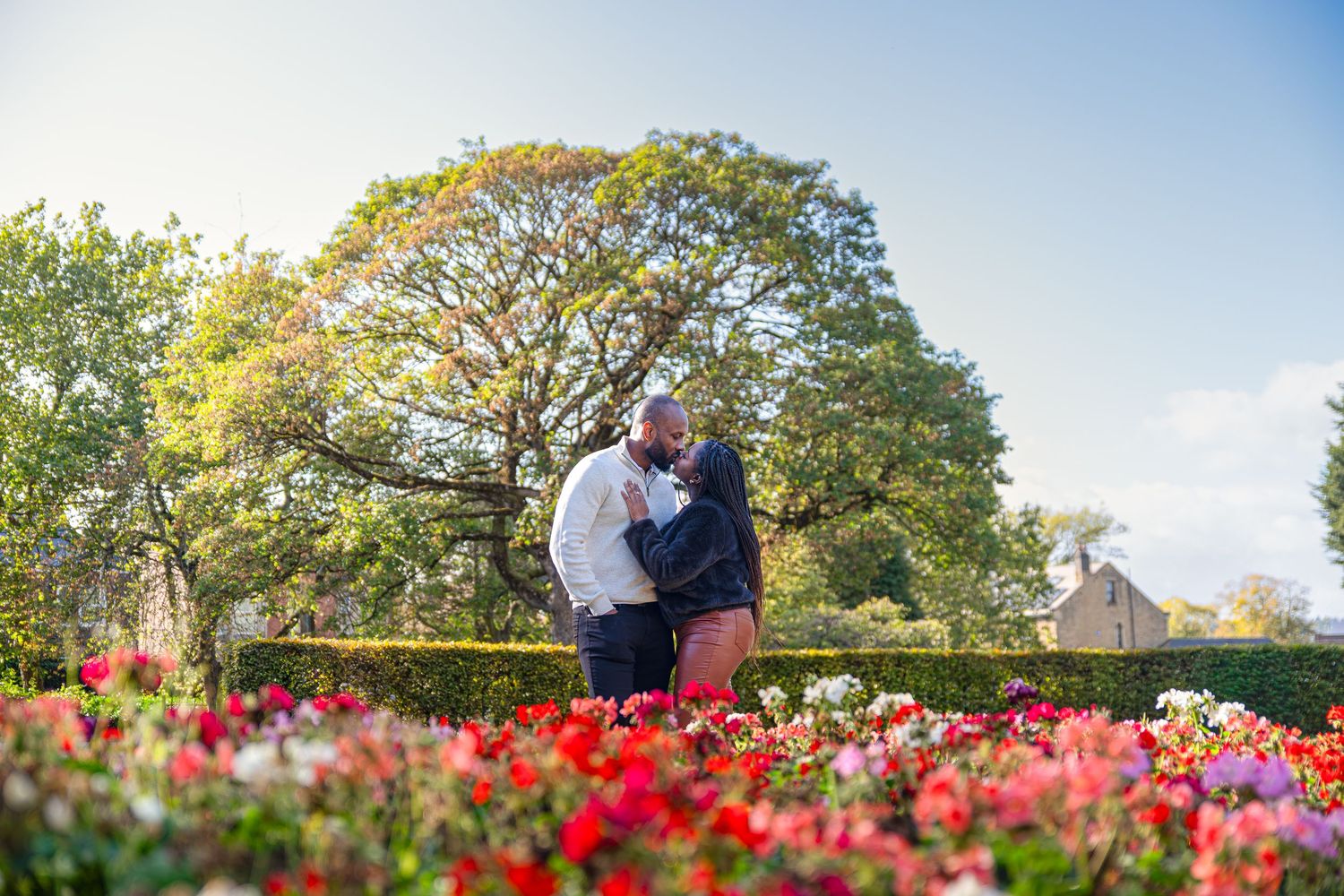 Couple sharing a romantic kiss in a vibrant garden with colorful flowers and lush green trees on a sunny day.