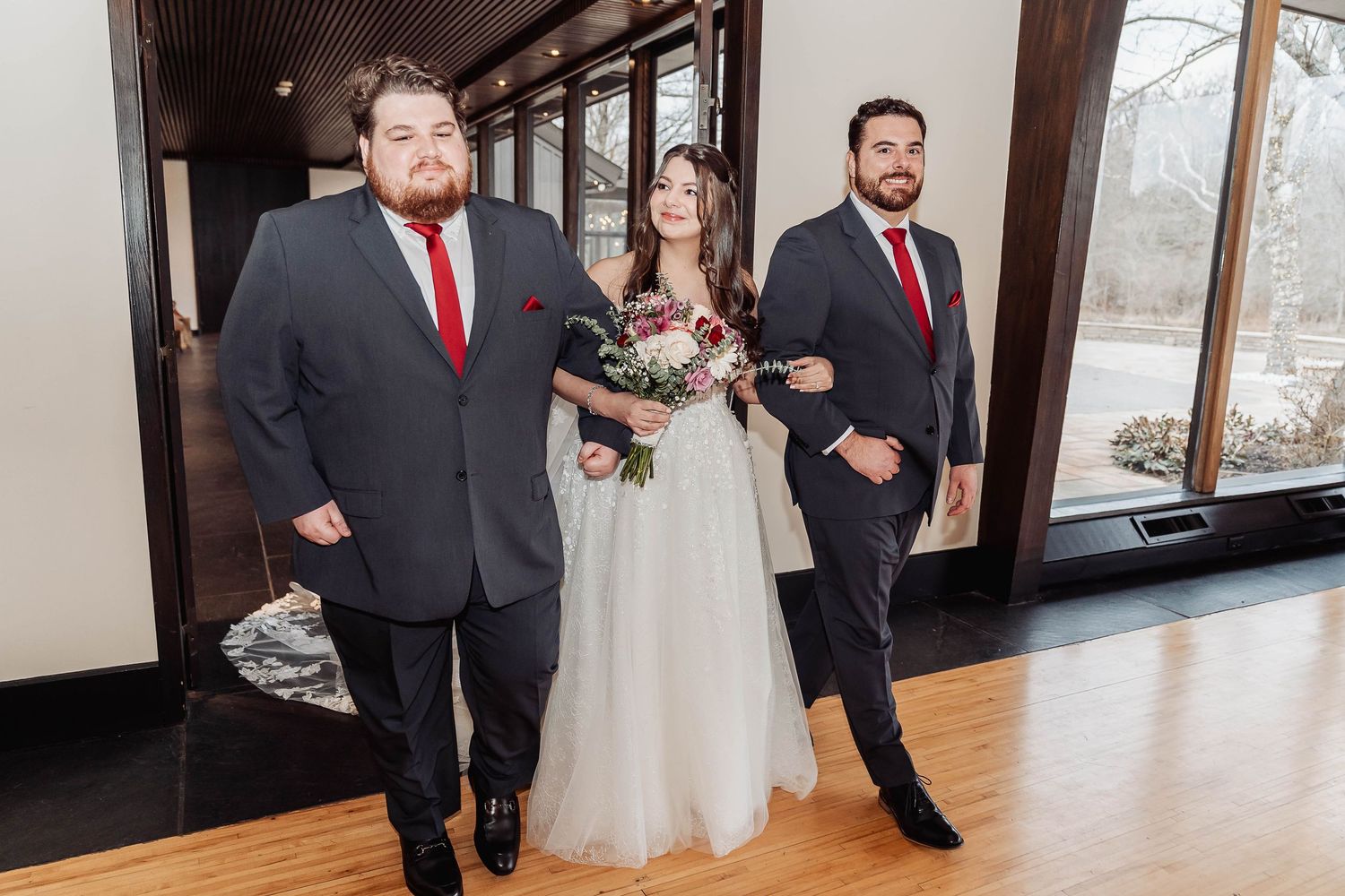 Bride in white gown with two groomsmen in dark suits and red ties at an elegant wedding venue.