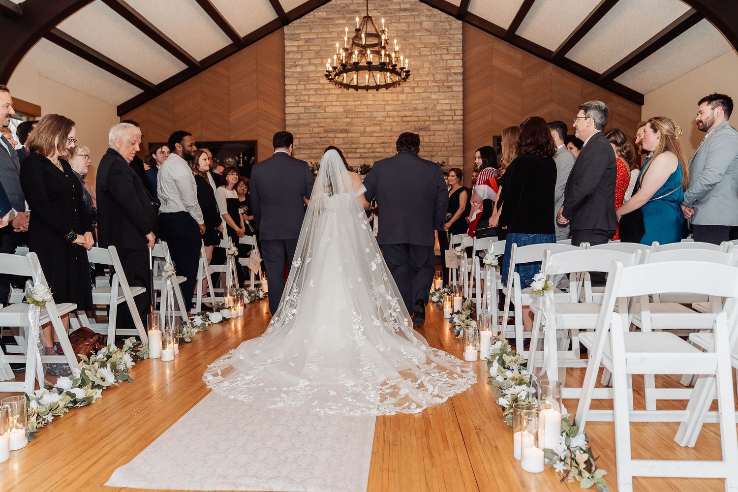 Bride and groom walk down the aisle at an elegant indoor wedding ceremony with white chairs and candlelit decor.