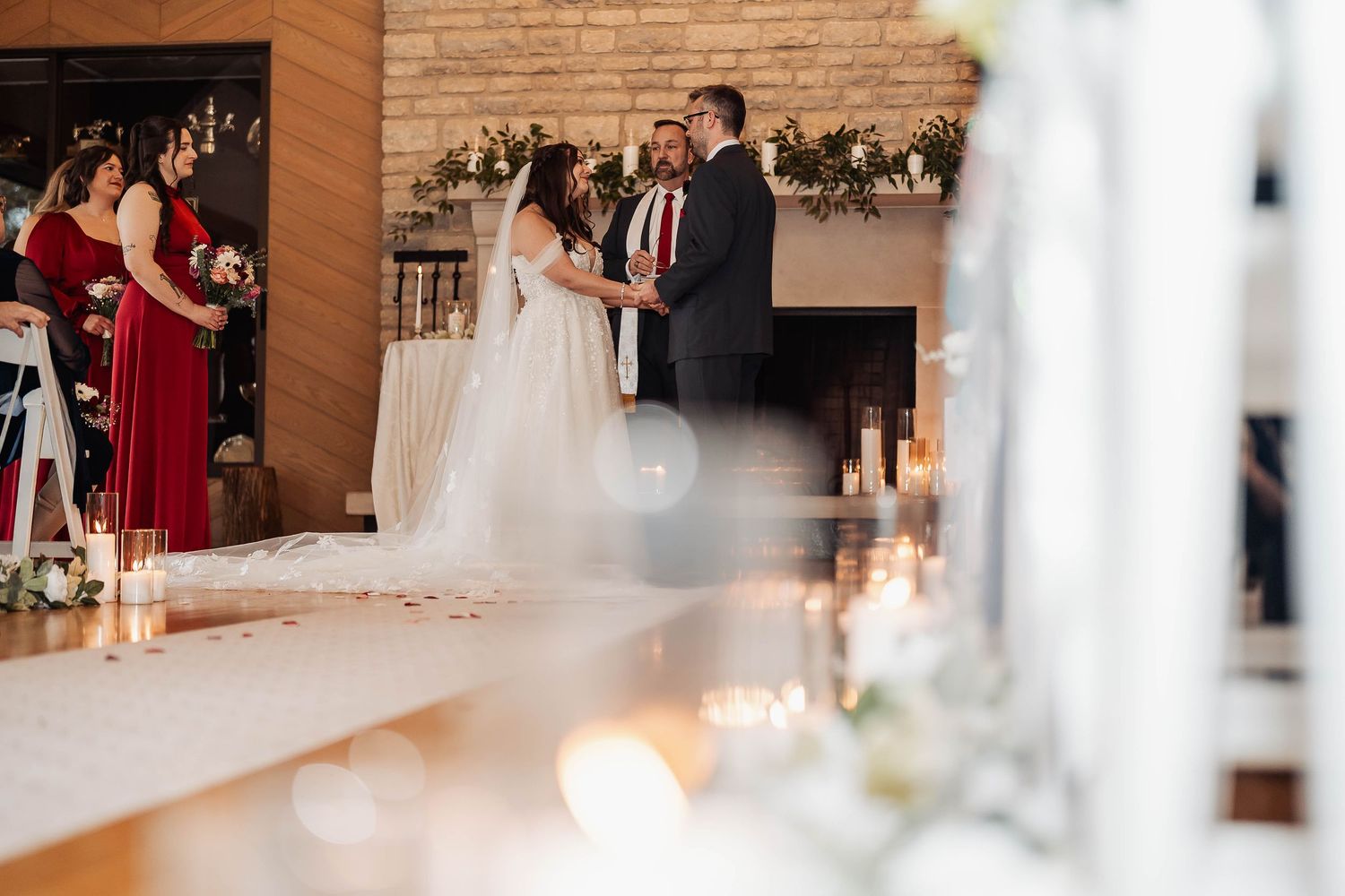 Couple exchanging vows at elegant indoor wedding ceremony with candles, floral arrangements, and brick fireplace backdrop.