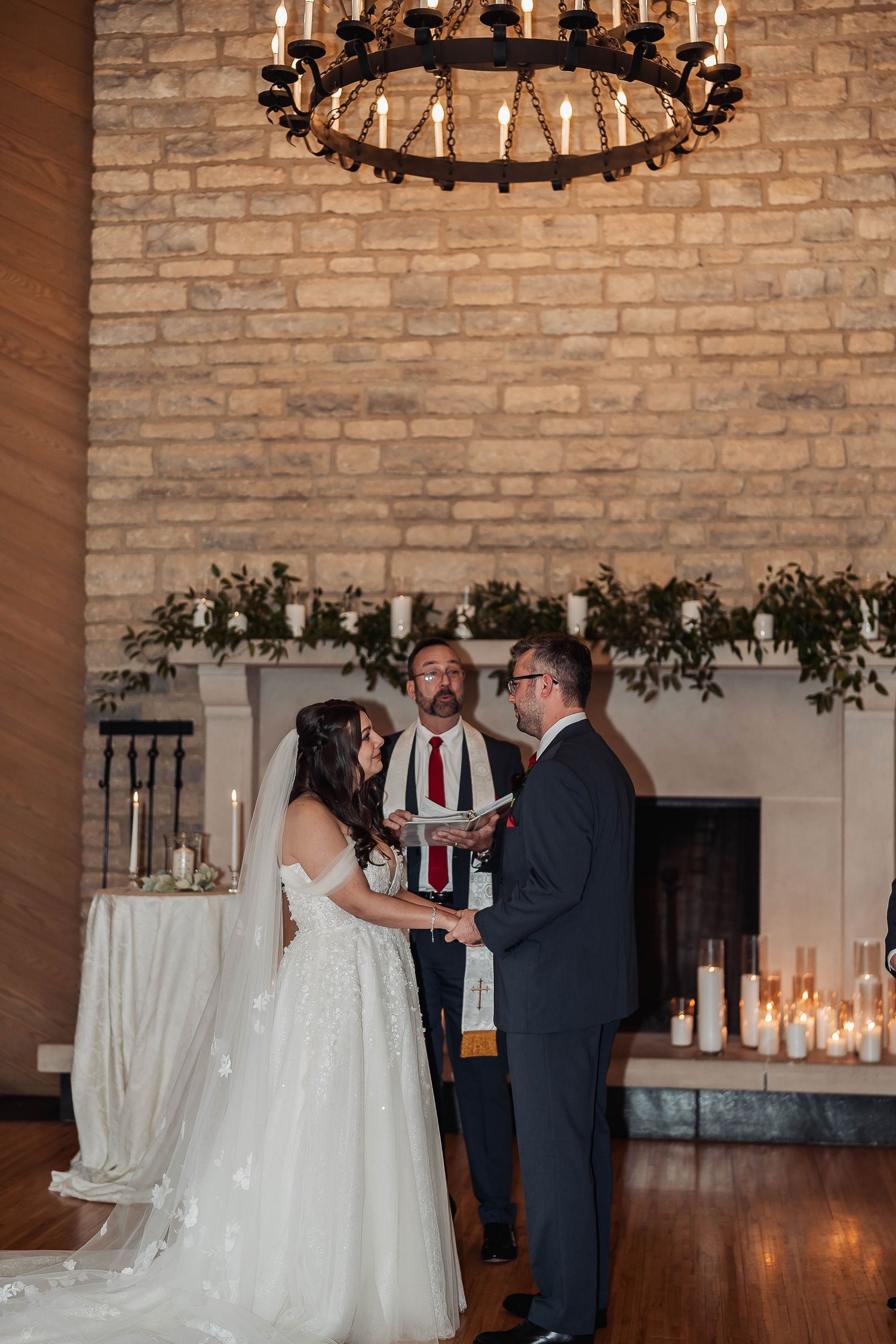 Bride and groom exchange vows before officiant at elegant indoor wedding ceremony with brick wall and candlelit fireplace.
