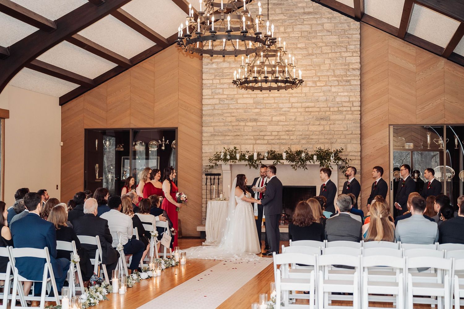 Elegant indoor wedding ceremony in a rustic venue with brick fireplace, chandelier, and guests seated in white chairs.