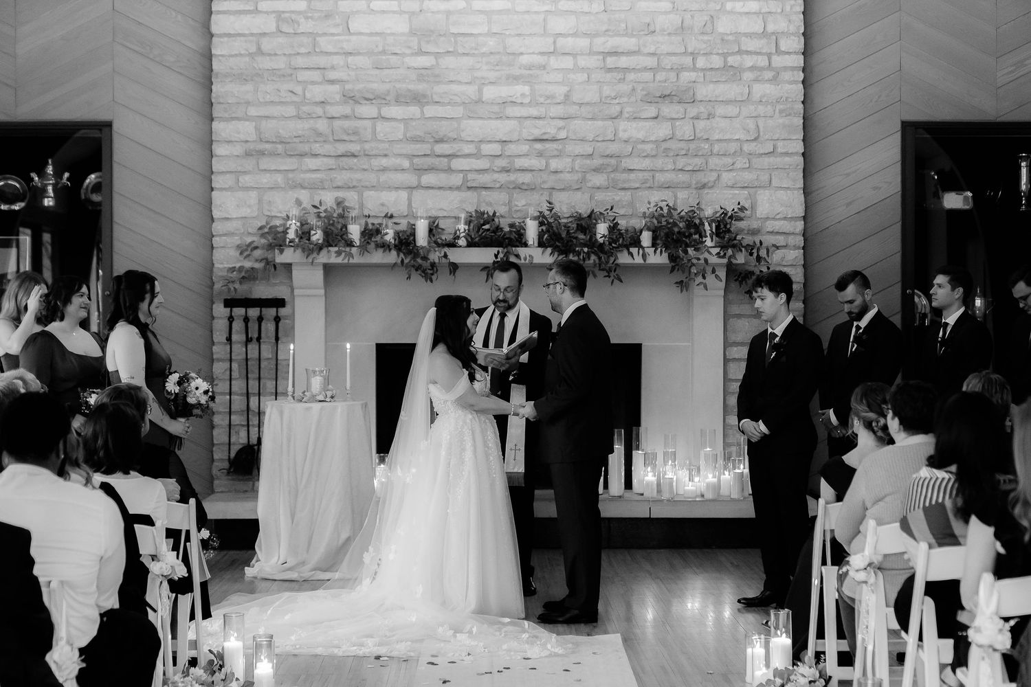 Black and white wedding ceremony photo with bride and groom at altar before fireplace, surrounded by guests and candles.
