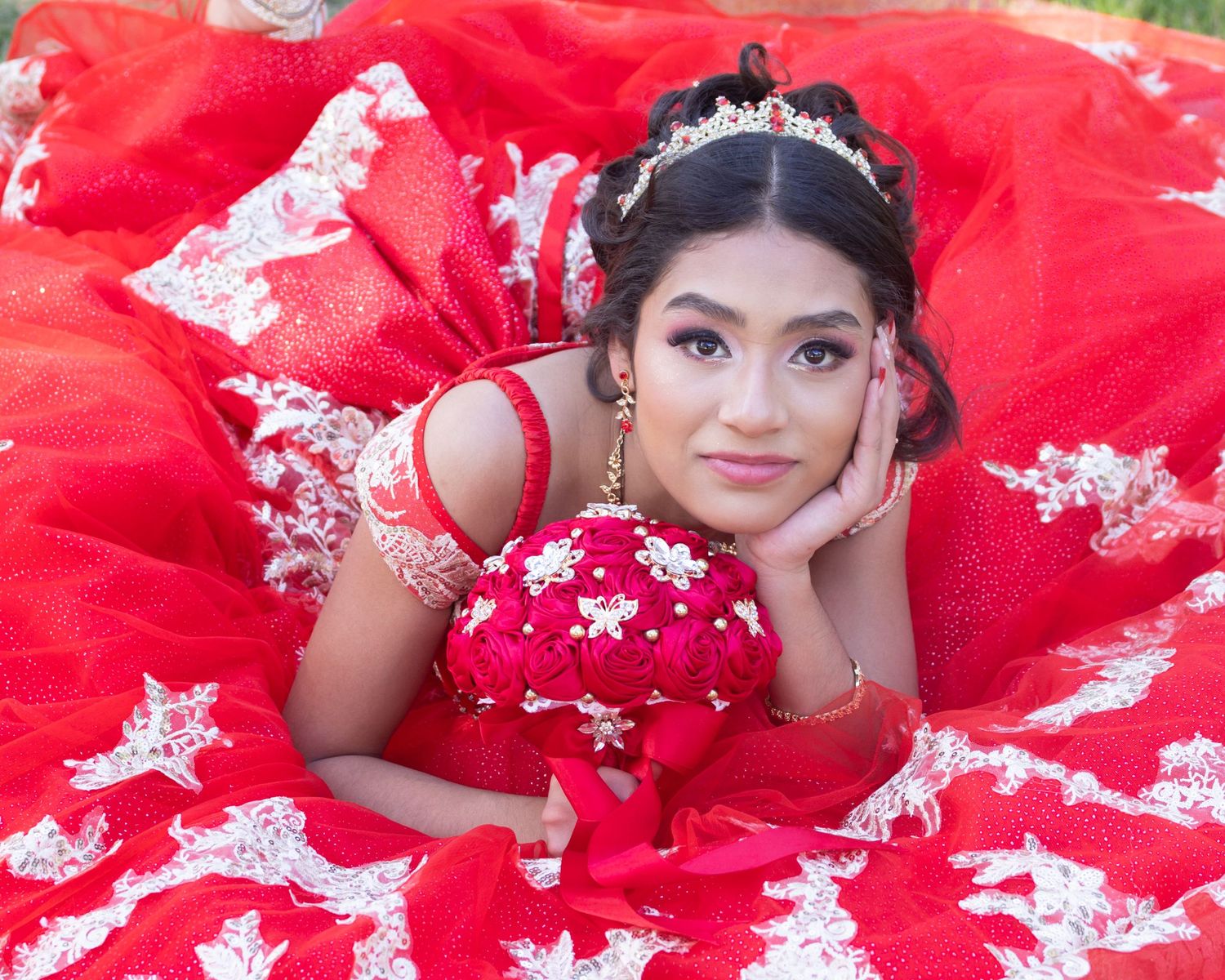 Young woman in red quinceañera gown with lace details, tiara, and floral bouquet, posing gracefully outdoors.