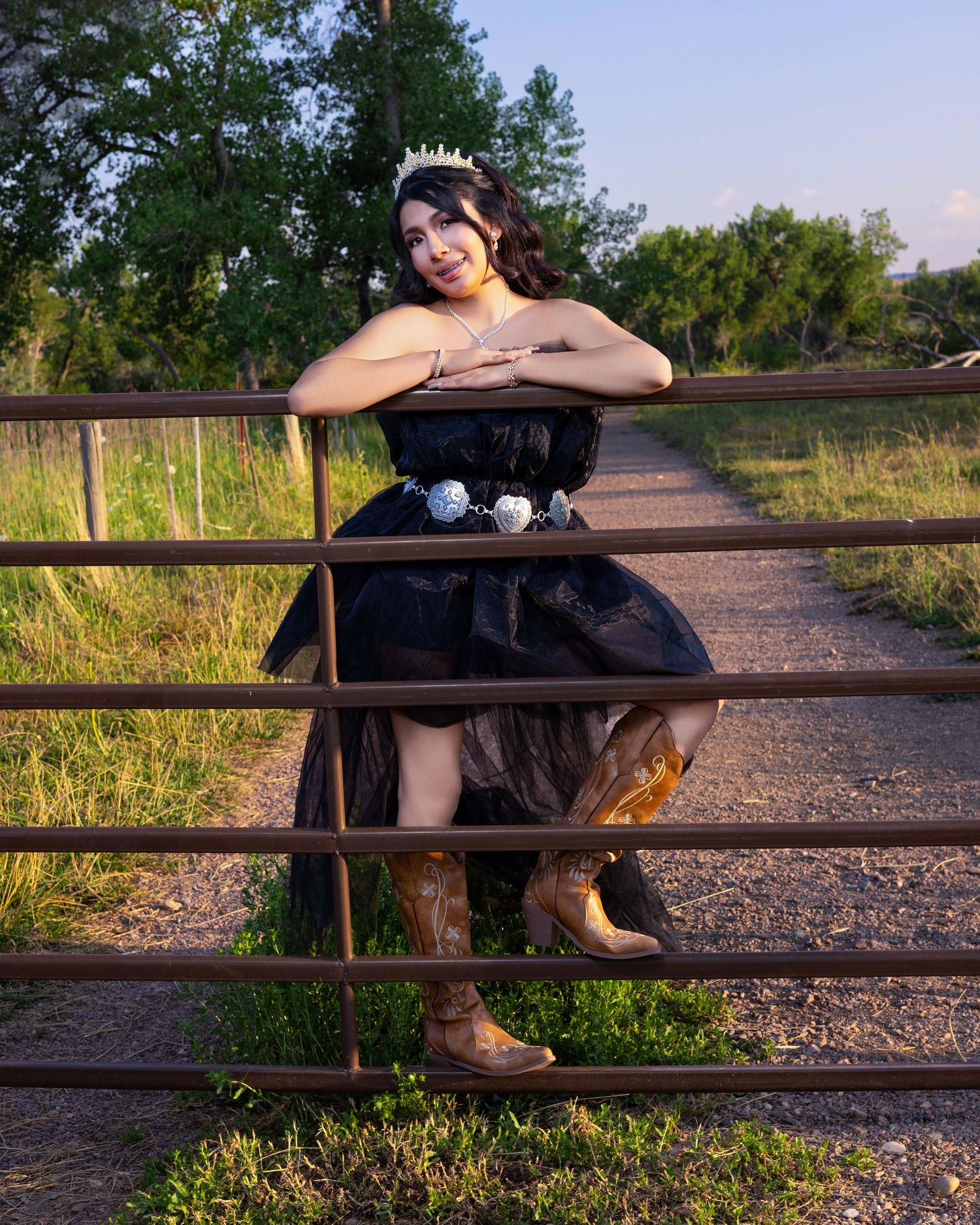 Young Latina woman in black tulle quinceañera gown with gold tiara and silver belt poses against tree bark.