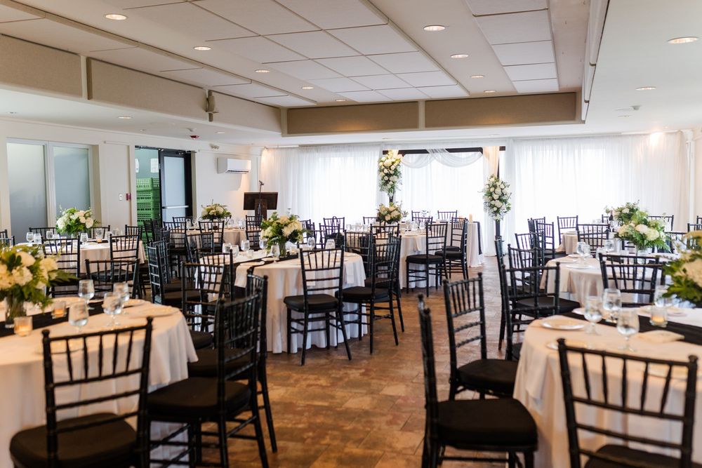 Elegant wedding reception hall with round tables, white linens, black Chiavari chairs, and floral centerpieces in a bright venue.