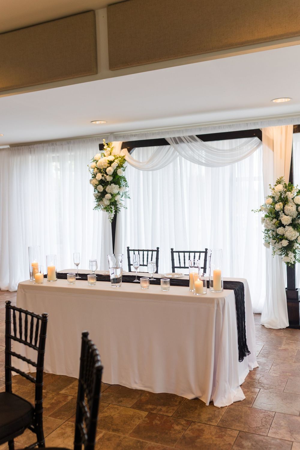 Elegant wedding sweetheart table with white draping, black chairs, tall floral arrangements, and candles in a banquet hall.